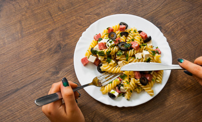 woman hand holding fork and knife with pasta fusilli salad with salami, cheese and olive in white plate on a wooden background. with copy space. top view