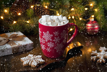  Close-up of a mug with a hot drink, marshmallow and gingerbread. Christmas decor with a bright garland and fir tree. Selective focus, shallow depth of field, background