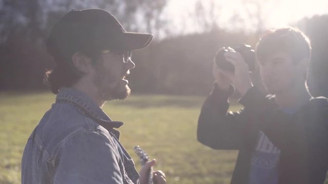 Shooting into the sun, a tight shot of teen male filmmaker filming a music video with a male guitar player and singer wearing a jean jacket and ball cap in the country.