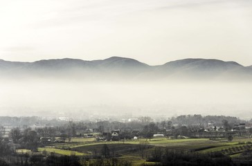 Fototapeta premium foggy rural landscape of village with green fields and mountains hill top