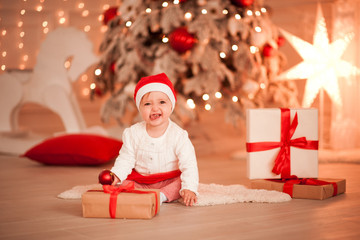 Fototapeta na wymiar Laughing baby girl 1 year old having fun with Christmas presents over Christmas lights in room. Looking at camera. Childhood.