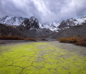 A dry lake bed pattern covered with green algae at the foot of the high mountain peaks with a snow caps under gloomy sky, Siberia, Altai, Shavla