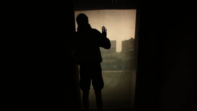 Silhouette man looking out the window with the light in the room. Footage. Silhouette of a young lonely man in the room. Concept of loneliness
