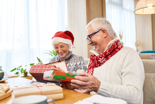 Portrait of loving senior couple unwrapping christmas presents and laughing happily sitting at dinner table with family home, copy space