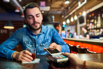 Portrait of drunk young man paying via smartphone buying drinks in bar, focus on payment terminal in foreground - obrazy, fototapety, plakaty