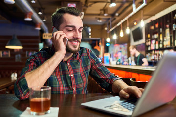 Portrait of modern young man working with laptop and speaking by phone at bar counter in beer pub