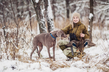 Female hunter in camouflage, armed with a rifle, standing in a snowy winter forest with duck prey
