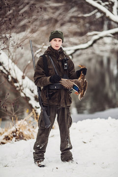 Male hunter in camouflage, armed with a rifle, standing in a snowy winter forest with duck prey