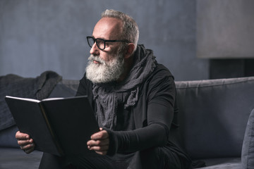 Side view pensive unshaven pensioner looking through volume while locating on comfortable sofa in...