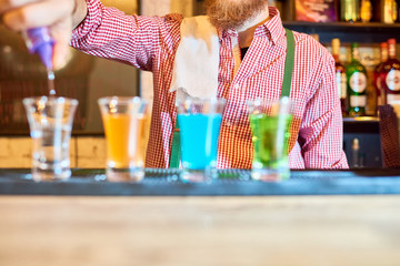 Mid section of unrecognizable bartender pouring alcohol shots at bar counter to little glasses of colorful liquid standing in row