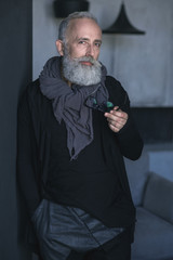 Portrait of serious unshaven mature male keeping glasses while leaning against wall. Fashion concept