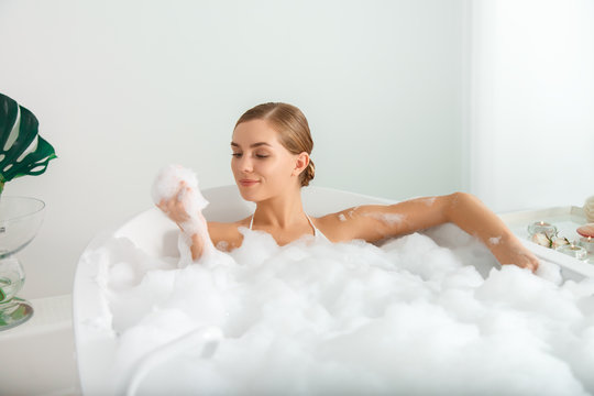 Cheerful young woman is lying in bathtub with relaxation. She is holding foam in hand and looking at it with interest. Purity and comfort concept