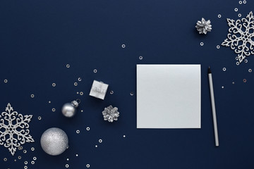 Silver blank Christmas card and a pencil with silvery New Year decorations, gift, snowflakes & sequins on dark blue background. Top view