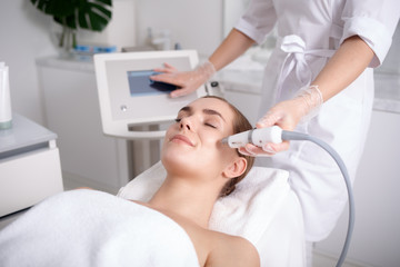 Side view of happy young woman getting cavitation rejuvenating skin treatment at spa. She is lying on massage table and smiling. Beautician is touching monitor screen while holding tool near female