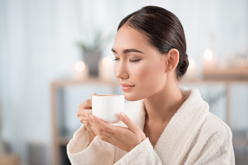 Great aroma. Gorgeous young asian lady is smelling fresh espresso with closed eyes and expressing satisfaction. She is resting in white bathrobe at beauty salon. Candles in background