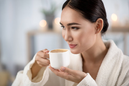 My favorite beverage. Attractive optimistic asian girl is drinking fresh coffee while relaxing in wellness center. She is looking aside with smile. Shelves with candles in background