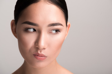 Close-up portrait of dissatisfied young asian woman is drawing faces in front of camera. She is standing and looking aside with discontent. Isolated background with copy space in the right side