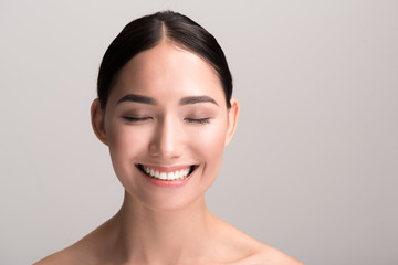 Joyful mood. Close-up portrait of positive young asian woman with clean and fresh skin is standing with closed eyes and feeling happy. Isolated background