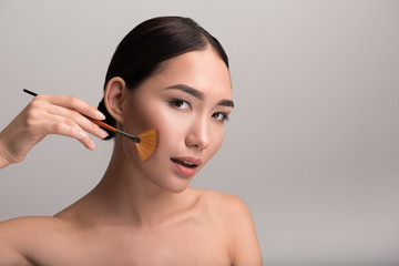 Flawless makeup. Portrait of tempted young naked asian lady is powdering her cheeks by professional brush. She is looking at camera seductively. Isolated background with copy space in the right side