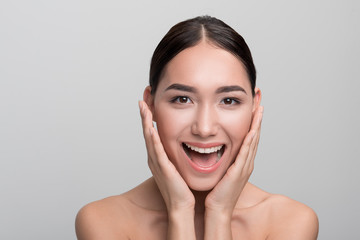 Sense of admiration. Portrait of cheerful naked young asian girl is touching her face while expressing happiness. She is looking at camera with wide smile while standing isolated. Skincare concept