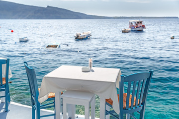 Fototapeta premium Served table with white tablecloth against blue water of Aegean sea on Santorini island resort in Greece, European famous and popular all year long resort.