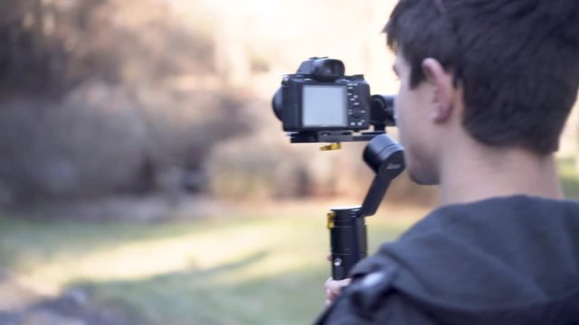 Teen filmmaker uses a gimbal to make a music video of a country rock star as the musician walks away from him down a gravel road.