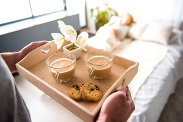 Top view close up of male hands holding tray with two cups of coffee, cookie and flowers. Pregnant woman is lying on bed on background