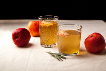 Two vintage glasses with apple cider on black background. Christmas beverages concept. Two red apples and rosemary sprig aside.  Warm backlight. Horizontal composition. Side view.
