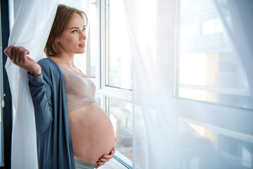 Side view of dreamful pregnant woman enjoying city view from window. She is touching her bare abdomen with love and smiling. Copy space