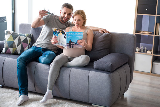 Planning future journey. Full length portrait of cheerful loving couple reading magazine about traveling while sitting on sofa at home. Man is holding plane toy while his pregnant wife is smiling