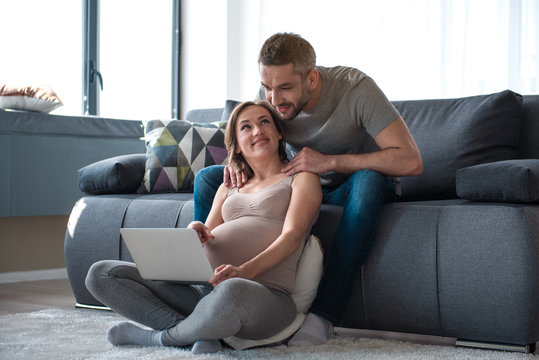 I want this one. Full length portrait of happy pregnant woman pointing on laptop screen and smiling. Her husband is looking there with interest while massaging female shoulders at home
