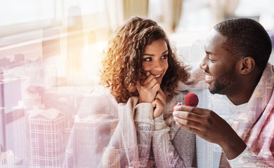 Be my wife. Close up of young pleasant couple sitting close to each other while holding a heart shaped box with wedding ring and expressing happiness