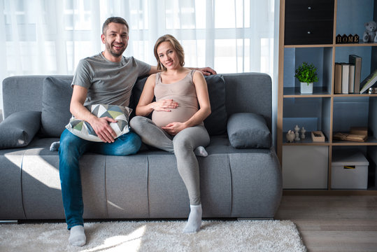 Happy family. Full length portrait of excited married couple sitting on sofa and smiling. Pregnant woman is touching her belly with joy