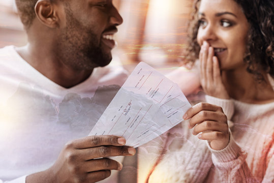 Surprise. Close up of young positive man presenting flight tickets and showing delight while his girlfriend covering her mouth with a hand and being pleased