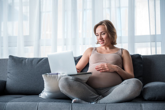Cheerful expectant mother is entertaining with portable computer at home. She is sitting on sofa with comfort and smiling. Full length portrait