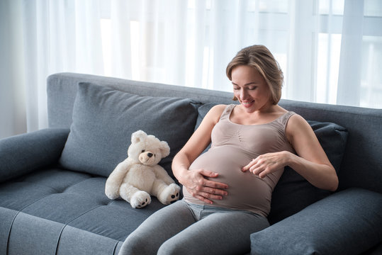 Excited pregnant woman is speaking with her unborn baby. She is touching tummy with interest and laughing while resting on couch. Copy space