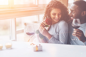 Favorite flower. Black man holding glass with red wine in left hand keeping smile on his face while embracing girlfriend