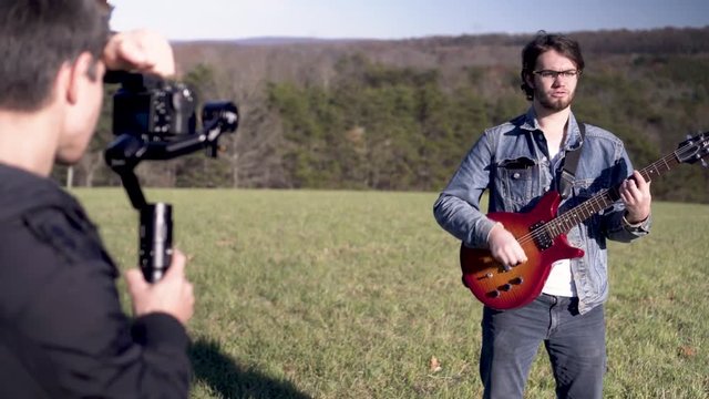 Young teen male caucasian filmmaker films a music video and orbits around his subject, a guitar musician and singer in a large grassy field under the bright sun.