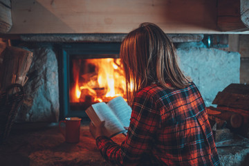 Woman reading a book by the fireplace. Young woman reading a book by the warm fireplace decorated for Christmas. Relaxed holiday evening concept.