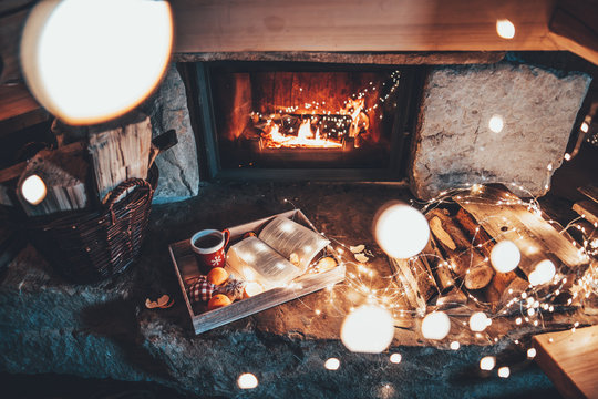 Warm cozy fireplace with real wood burning in it. Magical atmosphere. Cup of hot drink and book ready for evening relax. Cozy winter concept. Christmas and travel background with space for your text.