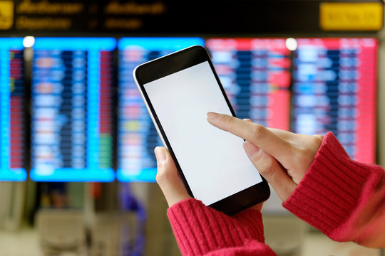 Mockup image of female hands holding black mobile phone with blank white screen over flight board in airport terminal.
