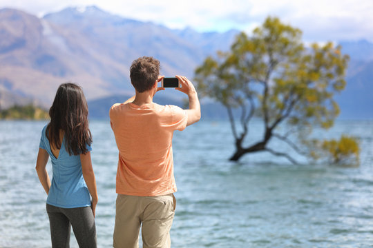 New Zealand tourists taking phone pictures of Wanaka Lone Tree at lake. People looking at view of famous touristic attraction in south island, Otago Region, NZ.