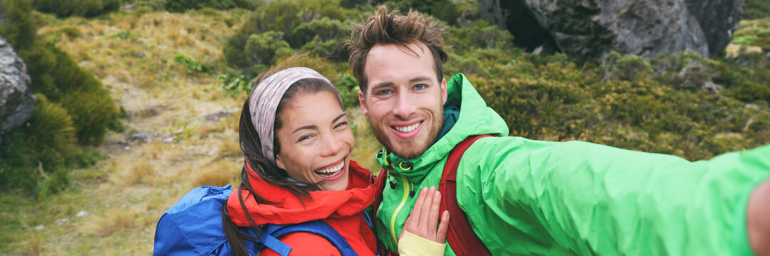 Travel selfie couple hikers taking smartphone picture on outdoor trail hike in outdoor nature. Active healthy happy people hiking panoramic banner. Multiracial young adults.