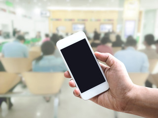 Hand holding mobile phone over blurred people sitting and waiting at bank counter or hospital.