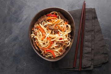 Close-up of noodles in a glass bowl with vegetables and beef in teriyaki sauce on a dark concrete background. Top view of Asian food, and wooden sticks. The concept of fast food
