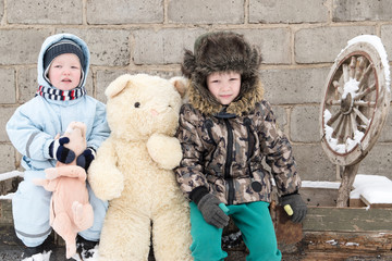 Two happy children in winter fashion clothes posing with a toy pig and a bear in the courtyard of a village house. First snow, family, tradition, holiday