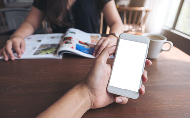 Mockup image of a man's hand holding and using white smart phone with blank screen on wooden table with woman reading magazine in background