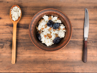 Clay bowl with cottage cheese, raisins and dried apricots next to the wooden spoon and knife. Dairy product cottage cheese in bowl on old table. Template for menu, poster or print design, top view.
