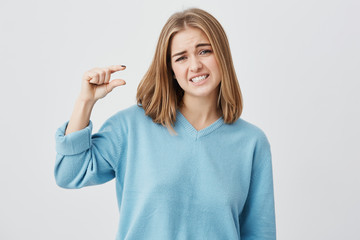 European young female wearing blue sweater with blonde hair showing something small in size with hands, gesturing, frowning brows and showing her teeth. Blonde demonstrating size of something.