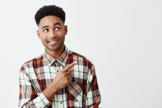 Portrait of joyful happy mature attractive black-skinned man with afro hairstyle in casual checkered shirt looking aside with raised eyebrows and smile, pointing with hand on white background. Copy